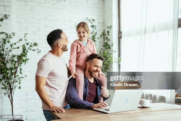 pareja gay adulta mediana con hija adoptiva en casa, trabajando en una computadora portátil y divirtiéndose con una niña - adopción fotografías e imágenes de stock