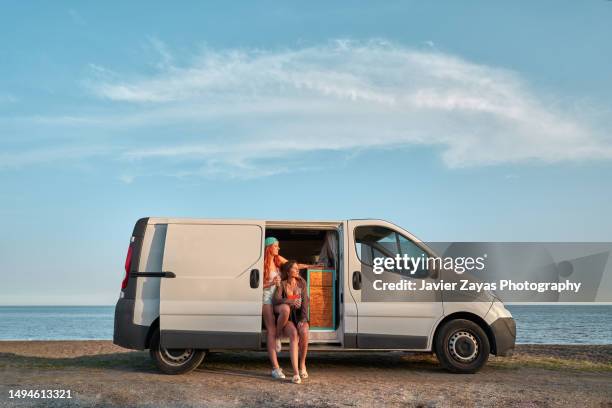 two female friends enjoying the sunset in camper van on the beach - lieferwagen stock-fotos und bilder