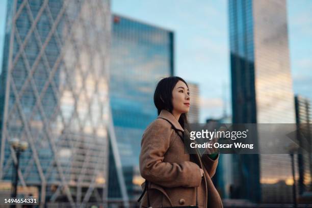 young asian business woman using smartphone standing against modern financial buildings in the city - town center stock pictures, royalty-free photos & images