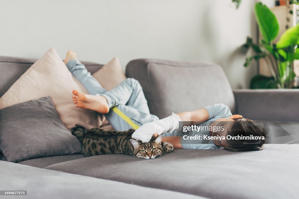 A boy with a broken arm plays with a cat at home