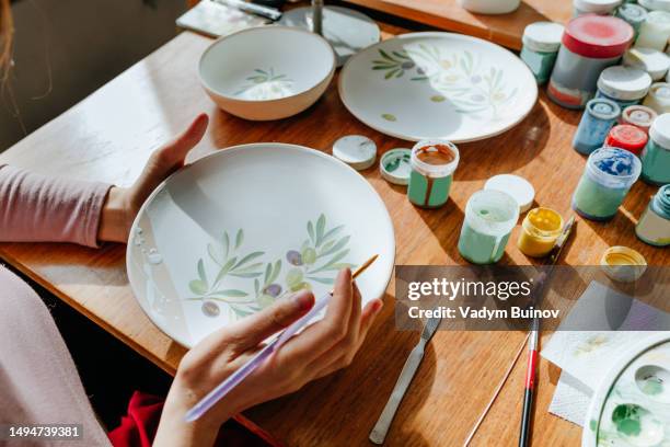 woman painting olive branch on clay plates with paintbrushes in a pottery workshop - serviesgoed stockfoto's en -beelden