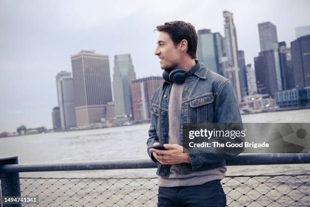 handsome man standing against city skyline holding smart phone - veste en jean photos et images de collection