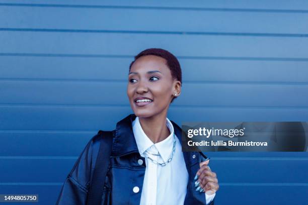 good-looking female tourist of african ethnicity in black leather jacket standing in front of blue wall holding jacket side with one hand - offener kragen stock-fotos und bilder