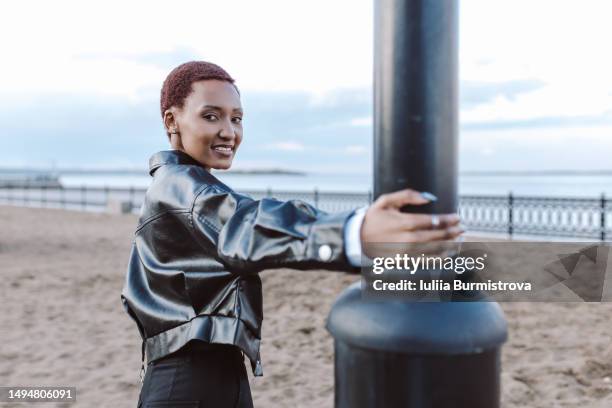 multiracial female traveler with short dark hai standing by street light post holding on to it on sandy beach - variable schärfentiefe stadt stock-fotos und bilder