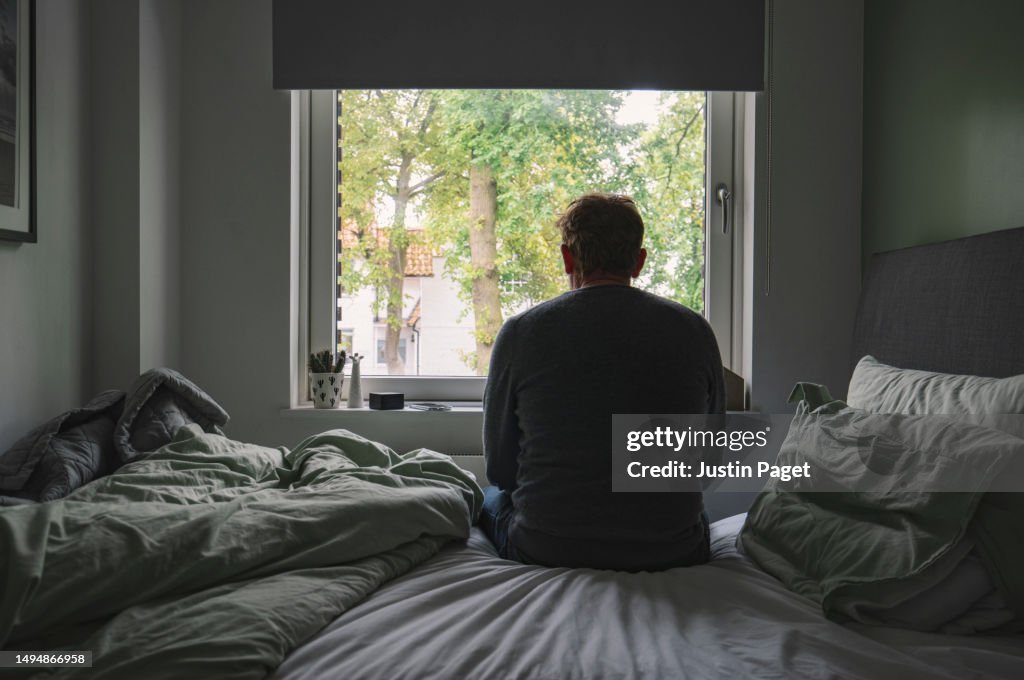 Rear view of an unrecognisable mature man sitting on his bed looking out of the window - negative emotion