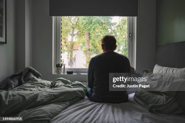 rear view of an unrecognisable mature man sitting on his bed looking out of the window - negative emotion - older men stock pictures, royalty-free photos & images