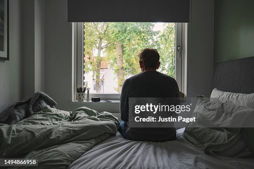 Rear view of an unrecognisable mature man sitting on his bed looking out of the window - negative emotion Rear view of an unrecognisable mature man sitting on his bed looking out of the window - negative emotion