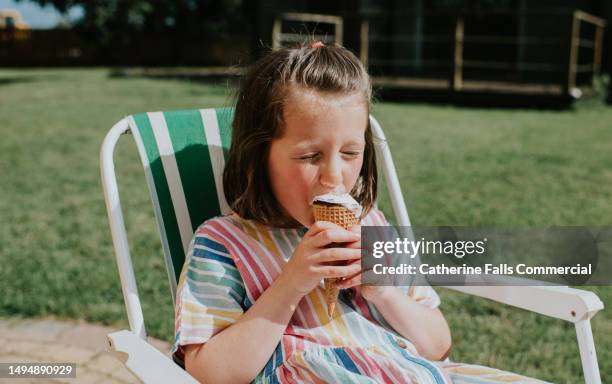 a little girl sits on a folding chair in a domestic garden, on a hot day, and eats an ice cream - hittegolf stockfoto's en -beelden