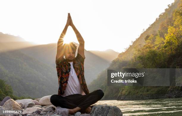 young man meditating on rocks at lakeshore during vacation - yoga day stock pictures, royalty-free photos & images