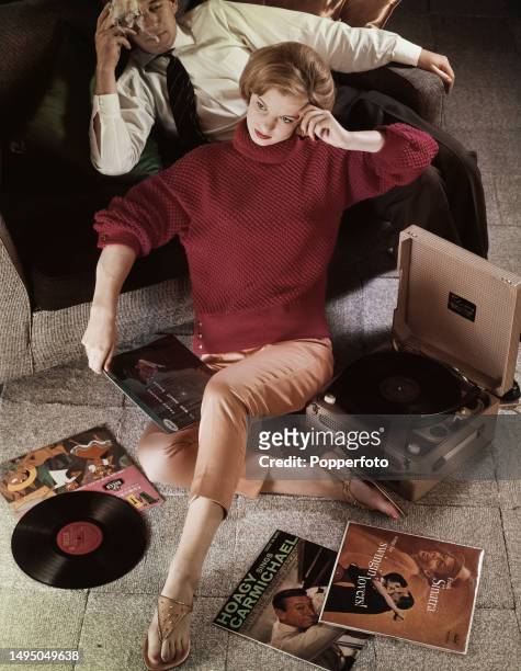 Posed studio portrait of a female fashion model wearing a red knitted lounging sweater and pale orange capri pants, she sits on the floor in front of...