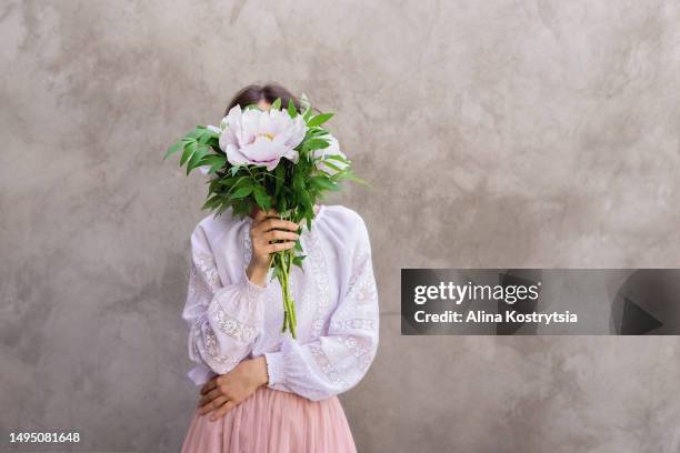 woman in white embroidered dress holds bouquet of peonies in front of her face - gesicht verstecken stock-fotos und bilder