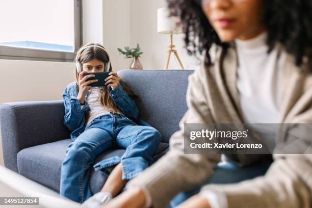 teen girl playing video games on mobile phone while sitting with her mother in the sofa at home. the woman is working on laptop computer. - addiction mobile and laptop stockfoto's en -beelden