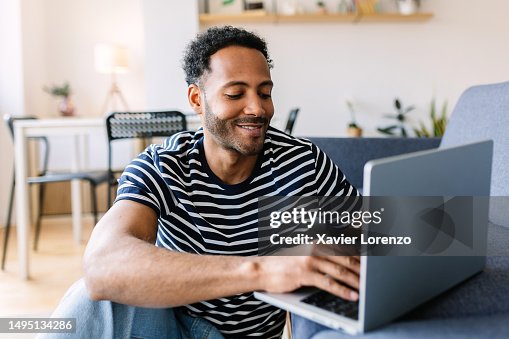 Happy young adult multiracial man using laptop computer while resting on sofa at home. Happy young adult multiracial man using laptop computer while resting on sofa at home.