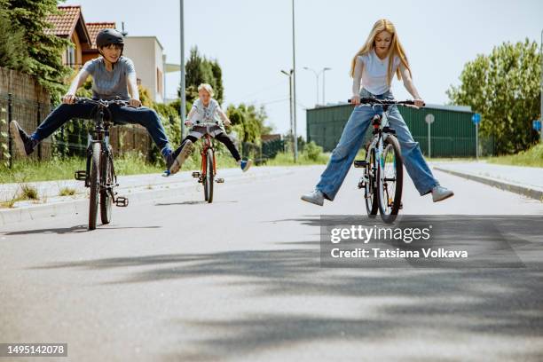 three children riding bikes on suburb street lifting their legs up from pedals - acrobazia foto e immagini stock