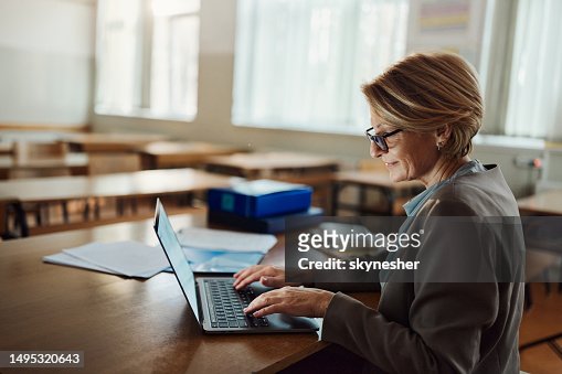 Smiling female teacher working on laptop in the classroom.
