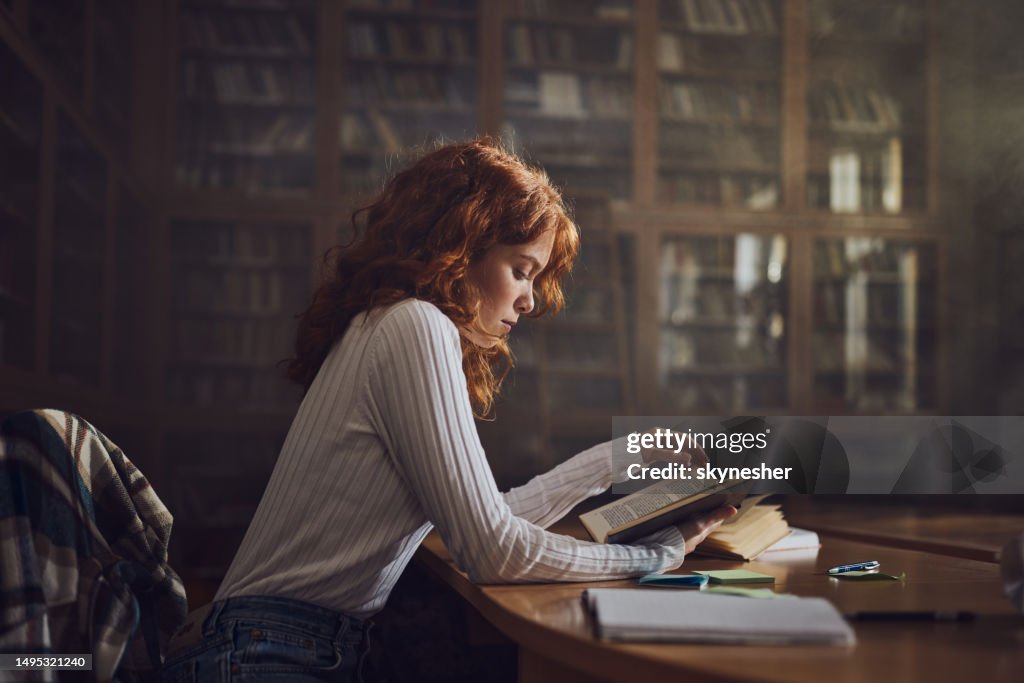 Redhead female student reading a book in library.