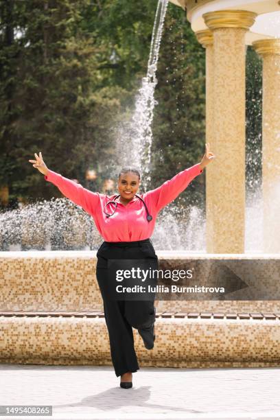 attractive female therapist of african ethnicity standing in front of fountain with arms raised smiling - on one leg stock pictures, royalty-free photos & images