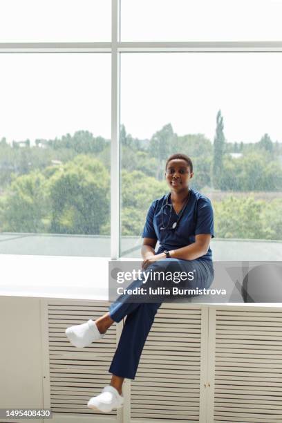 good-looking female nurse of african ethnicity wearing hospital gown, crocs and stethoscope sitting on window sill - nurse thinking stock pictures, royalty-free photos & images