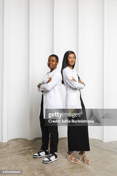 two young women with black hair in white lab coats standing back to back in front of white wall - showus doctor stock pictures, royalty-free photos & images