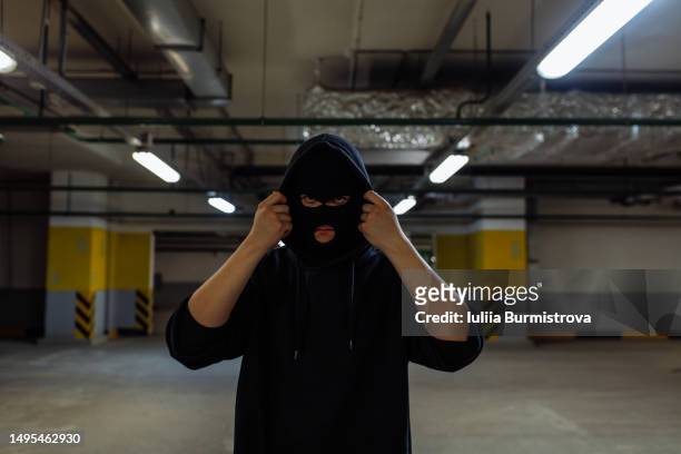 sinister-looking young man wearing face mask and hooded sweatshirt standing in empty parking garage - mugger stock pictures, royalty-free photos & images