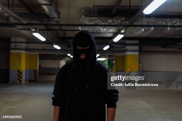 portrait of hooded man wearing black face mask standing in empty underground parking garage - mugger stock pictures, royalty-free photos & images
