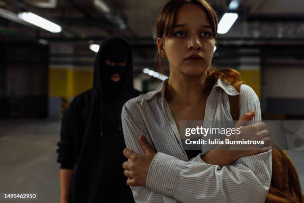 terrified young woman walking along underground parking lot while sinister man in mask following behind - mugger stock pictures, royalty-free photos & images