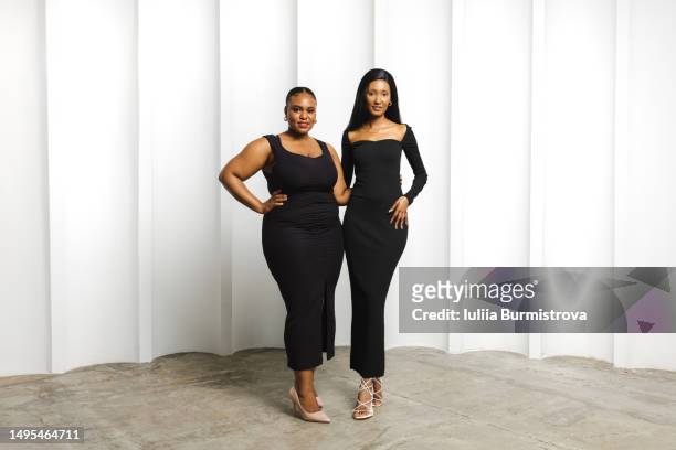 two attractive multiracial fashion models with black hair in long fitting black dresses standing close in front of white wall - form fitted dress stock pictures, royalty-free photos & images