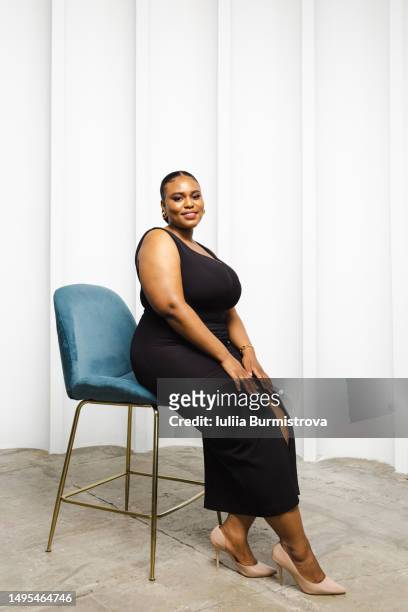 confident woman of african ethnicity with plump sensual body in long black dress sitting on high blue chair - fat women in high heels 個照片及圖片檔