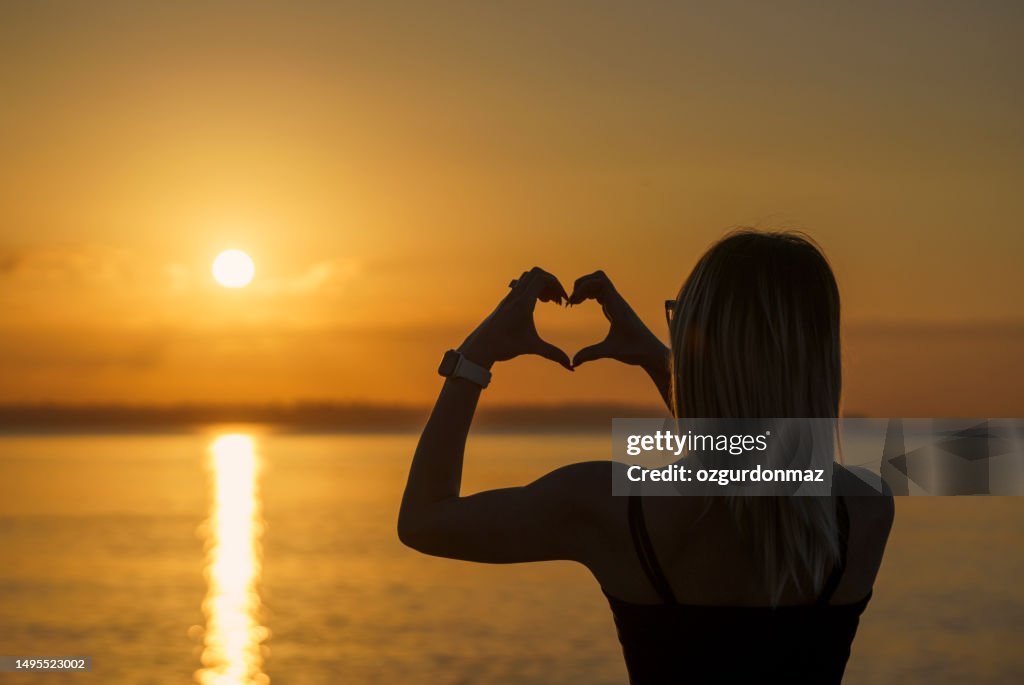 Woman's hands making a heart shape over a sunset landscape