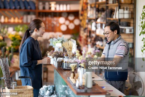 using a point of sale system (pos) can increase customer relationships and satisfaction in retail business. a male japanese shop owner or cashier talks with a customer and scans a price tag via a barcode reader to charge customers in a home decor store. - tag 2 stockfoto's en -beelden