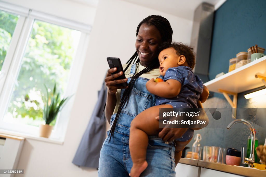 Mother of black ethnicity texting messages on smart phone while holding her baby son