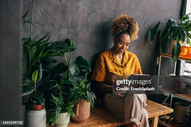 a happy beautiful blonde businesswoman using her tablet while sitting indoors surrounded by her houseplants - reading ipad stock pictures, royalty-free photos & images