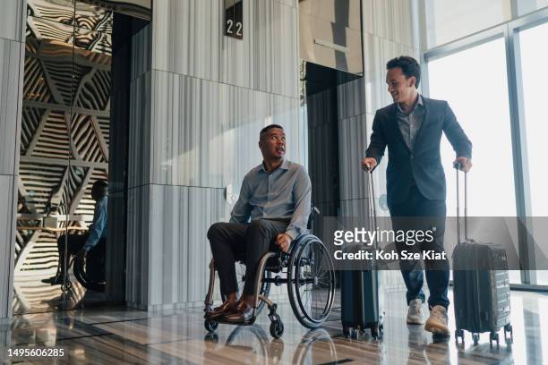 two malay businessmen on business travel one of whom is disabled wheeling their luggage out of the elevator lobby - one friend helping two other imagens e fotografias de stock