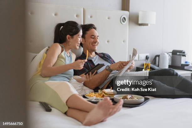 holiday pleasures - asian couple enjoying in room service and breakfast in bed - read and newspaper and bed stock pictures, royalty-free photos & images