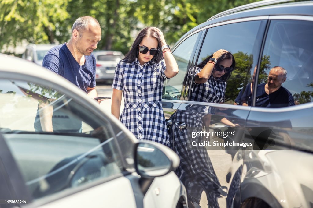 Woman who scratched a car on a parking lot holds her head and looks at the dent with the male owner of the damaged car.