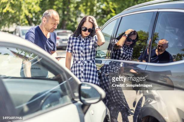 woman who scratched a car on a parking lot holds her head and looks at the dent with the male owner of the damaged car. - abollado fotografías e imágenes de stock