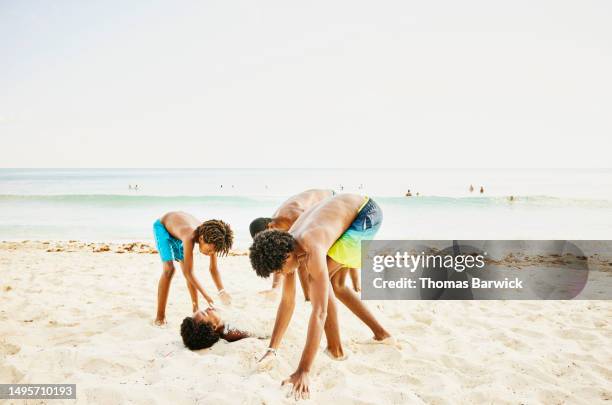 wide shot of boys burying younger brother in sand on tropical beach - bury fotografías e imágenes de stock