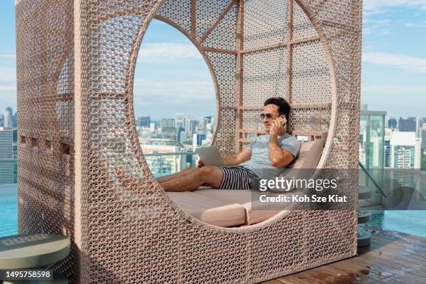 asian man on holiday relaxing by the poolside and occasionally checking on work - singapore travel stock pictures, royalty-free photos & images
