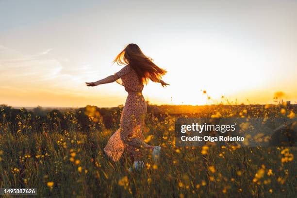 young woman in long dress dances with joy in a rapeseed yellow field at sunset - lente stockfoto's en -beelden