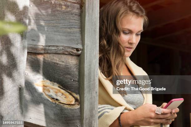 young woman texting through smart phone while standing by wooden wall - scialle foto e immagini stock