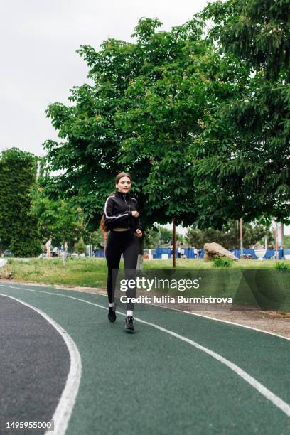 sporty young female sports person in black jogging suit running at sports stadium - estadio de atletismo fotografías e imágenes de stock