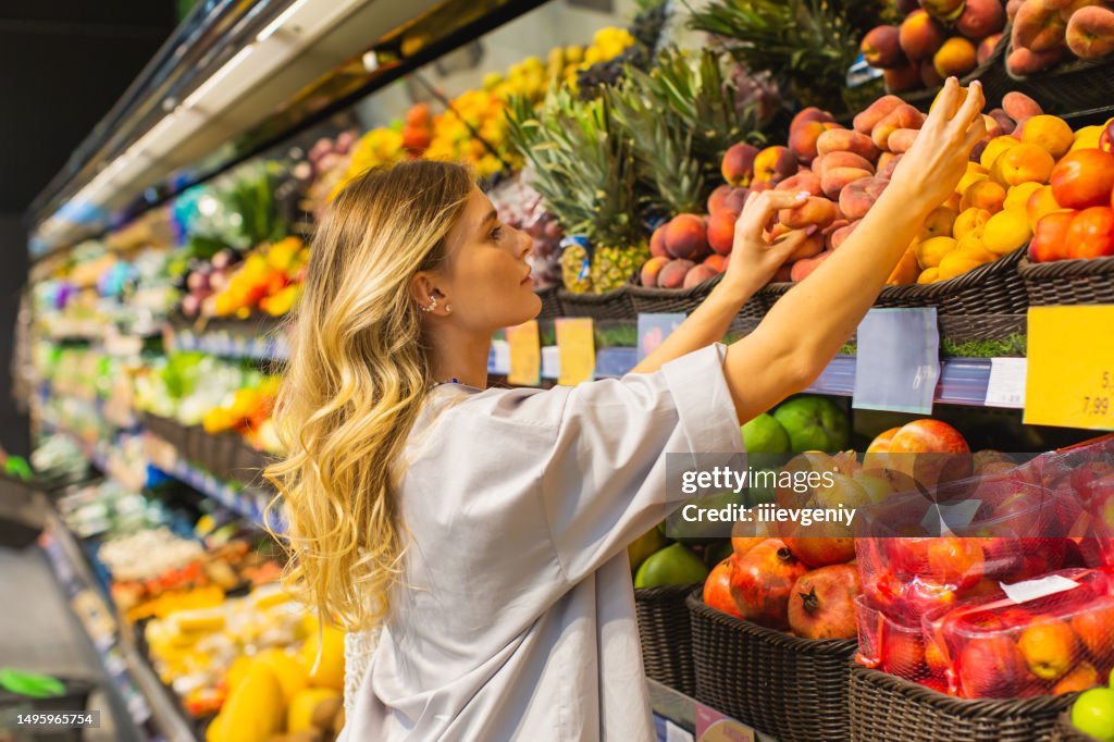 Junge blonde Frau wählt Pfirsich in der Obstabteilung. Herbstmarkt. Vegetarismus. Gesundes Essen. Wahl. Einkaufen. Wählen Sie Früchte nach Duft aus. Essen schnüffeln