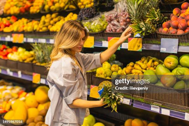 young blonde woman chooses pineapple in fruit section. autumn market. vegetarianism. healthy food. choice. shopping - tropical fruit stock pictures, royalty-free photos & images