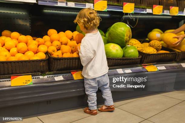 small blonde hair child boy in shop with orange in hand. fruit section. autumn market. vegetarianism. healthy food. choice. shopping - human back stock pictures, royalty-free photos & images