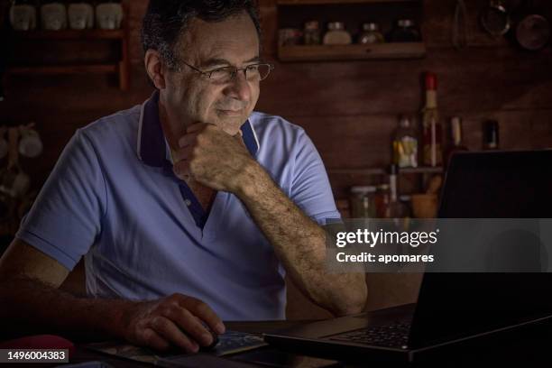 nahaufnahme von senior hispanic mann mit laptop in einer häuslichen küche home-office mit niedriger beleuchtung - close up phone on surface stock-fotos und bilder