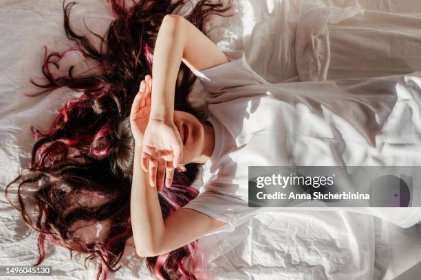 attractive young woman lies on bed with white textured fabric. charming brunette in a white t-shirt and beautiful long curly highlighted hair closes her eyes with her hands because of the bright sun. - cabello-teñido fotografías e imágenes de stock