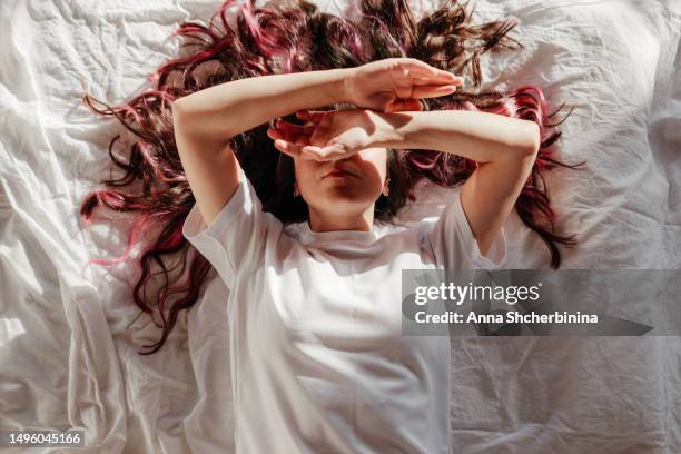 attractive young woman lies on bed with white textured fabric. charming brunette in a white t-shirt and beautiful long curly highlighted hair closes her eyes with her hands because of the bright sun. - thick white women ストックフォトと画像