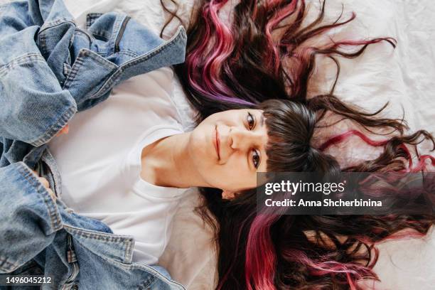 young woman with beautiful wavy highlighted hair, thick black bang and pink strands lies on white texture fabric. attractive smiling brunette girl in white t-shirt and denim jacket. - thick white women ストックフォトと画像