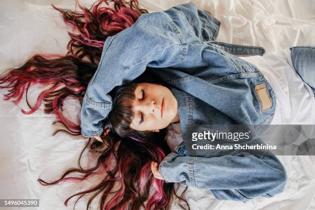 young woman with beautiful wavy highlighted hair, thick black bang and pink strands lies on white texture fabric. attractive smiling brunette girl in white t-shirt and denim jacket closed her eyes. - thick white women ストックフォトと画像