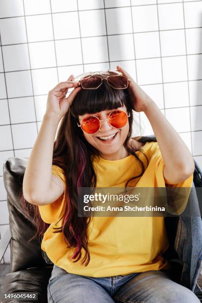 young attractive woman with beautiful long highlighted hair and round red sunglasses sits on black armchair and laughs. portrait of charming cute girl in yellow t-shirt and three pairs of glasses. - thick black woman 個照片及圖片檔